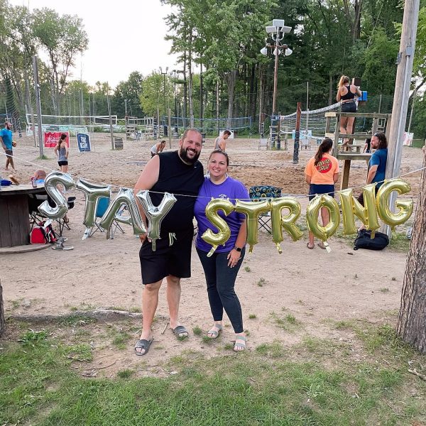 Man and woman with balloons spelling "Stay Strong" standing in front of beach volleyball courts