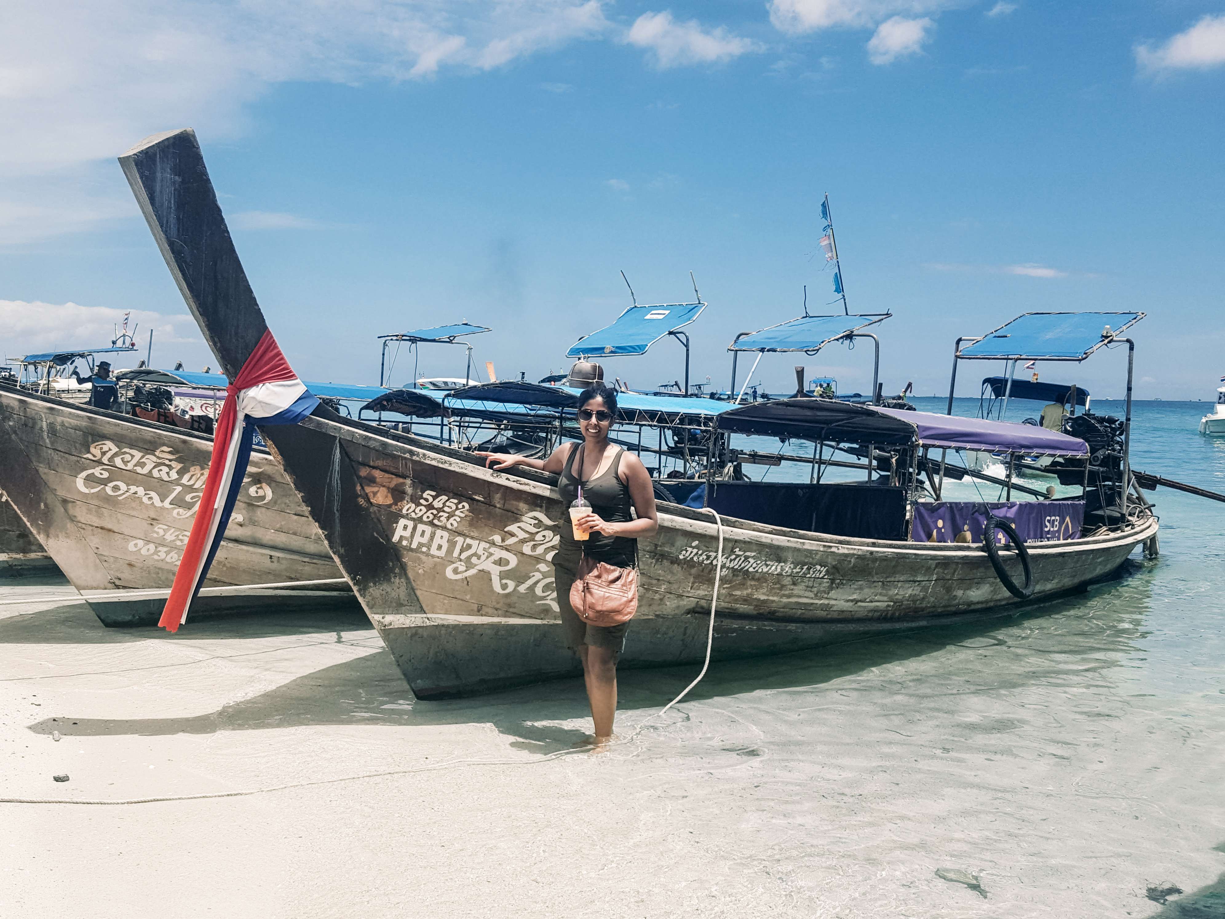 Woman poses on the shore in front of two brown sailboats with white text, with a blue body of water in the background.