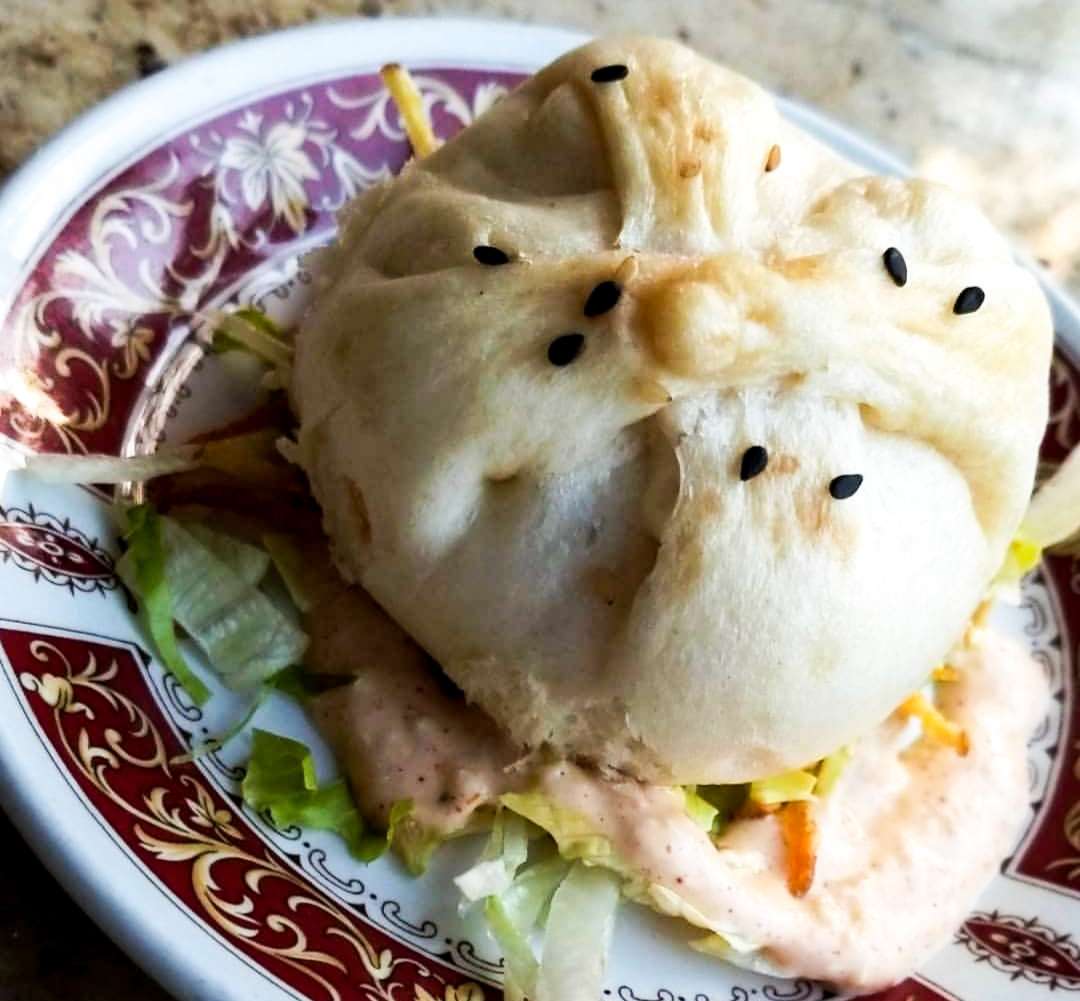 a bao which is a round bun with black sesame seeds on it, sitting on top of an light peach sauce and lettuce in a small round plate. 