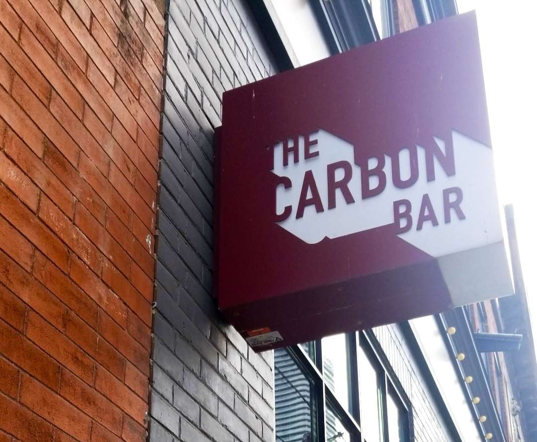 a square red and white sign with the name carbon bar on it, attached to a brick wall. 