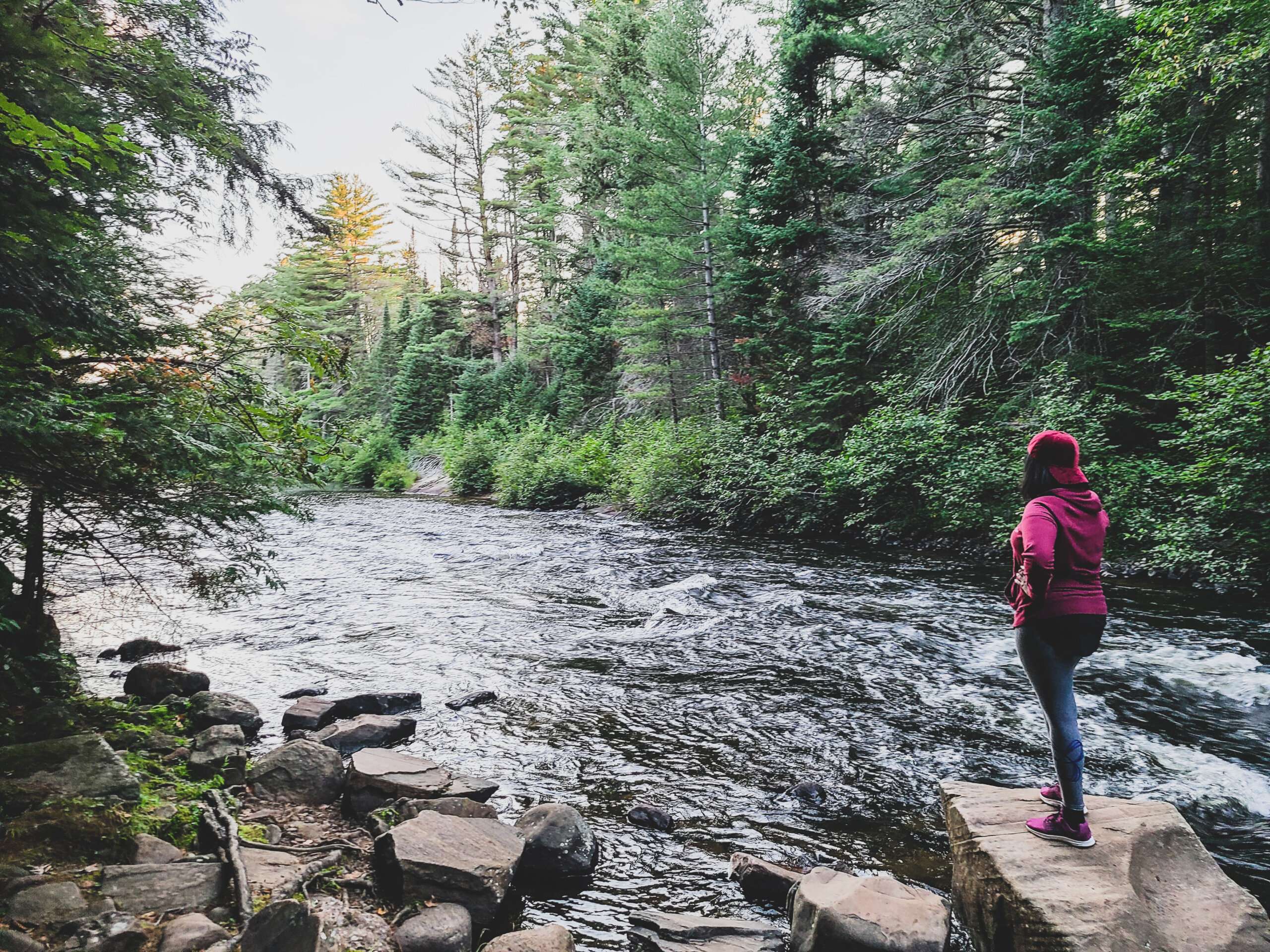 a women wearing a burgundy sweater with grey pants and a red cap standing on a rock looking out into water rapids surrounded by green trees