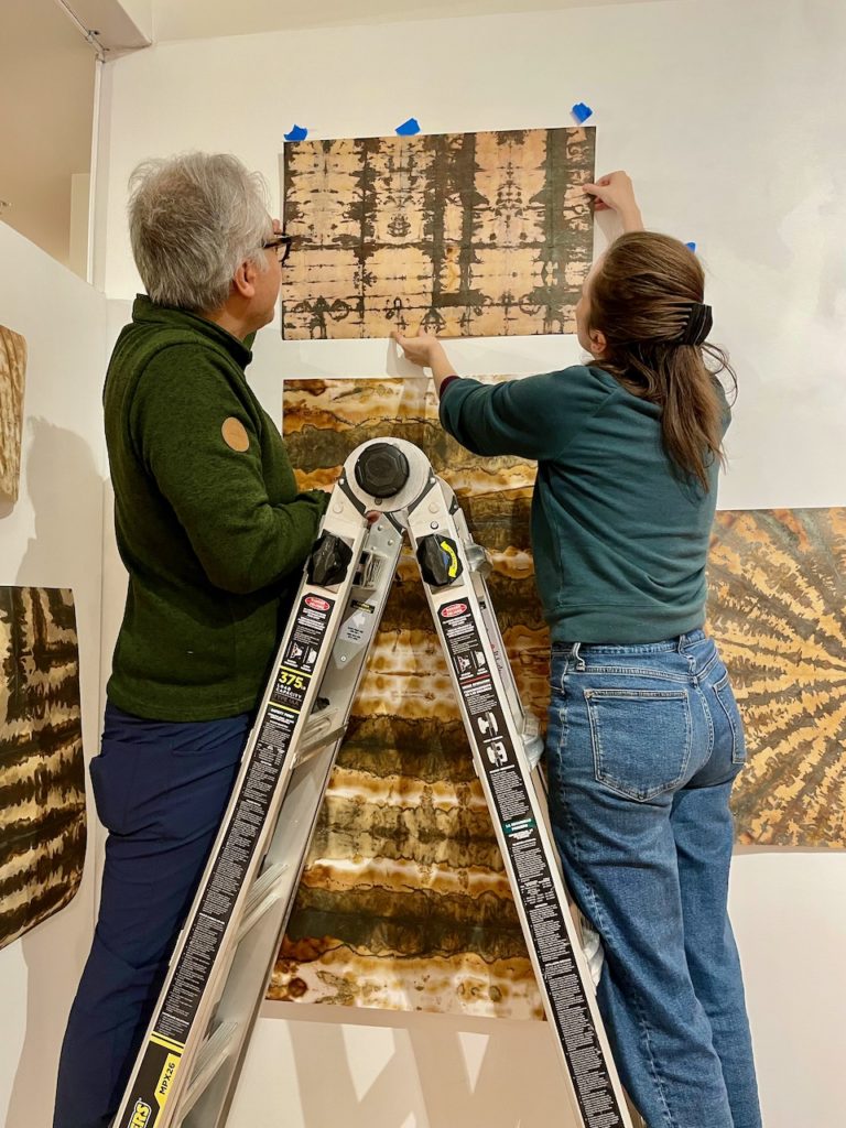 Larry and Betsy positioning a print about to be hung on the wall. they're each standing on one side of the same ladder