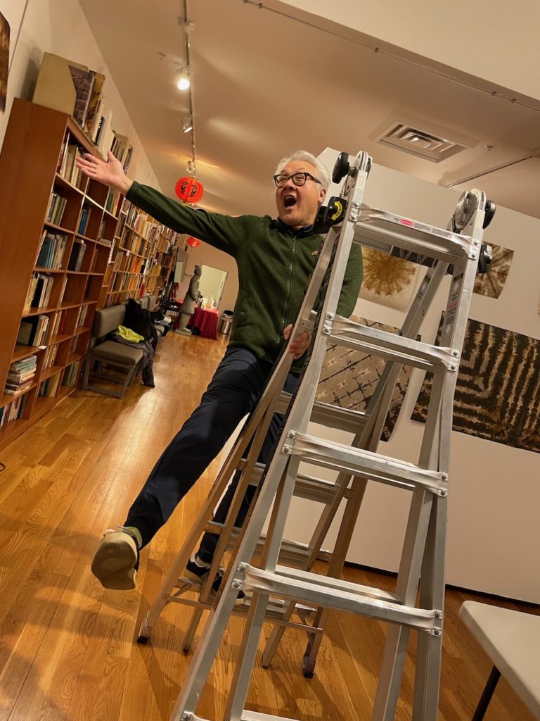 Larry Lee triumphantly throwing his right arm and leg out while balancing on a ladder with his left leg and arm