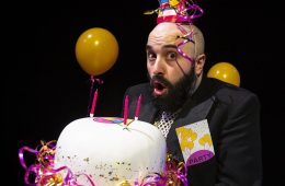 A man with a surprised expression is holding a birthday cake and wearing a brightly coloured party hat.