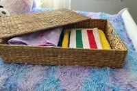 The Pottery Barn Savannah Handwoven Seagrass Lidded Underbed Basket partially open on top of a bed