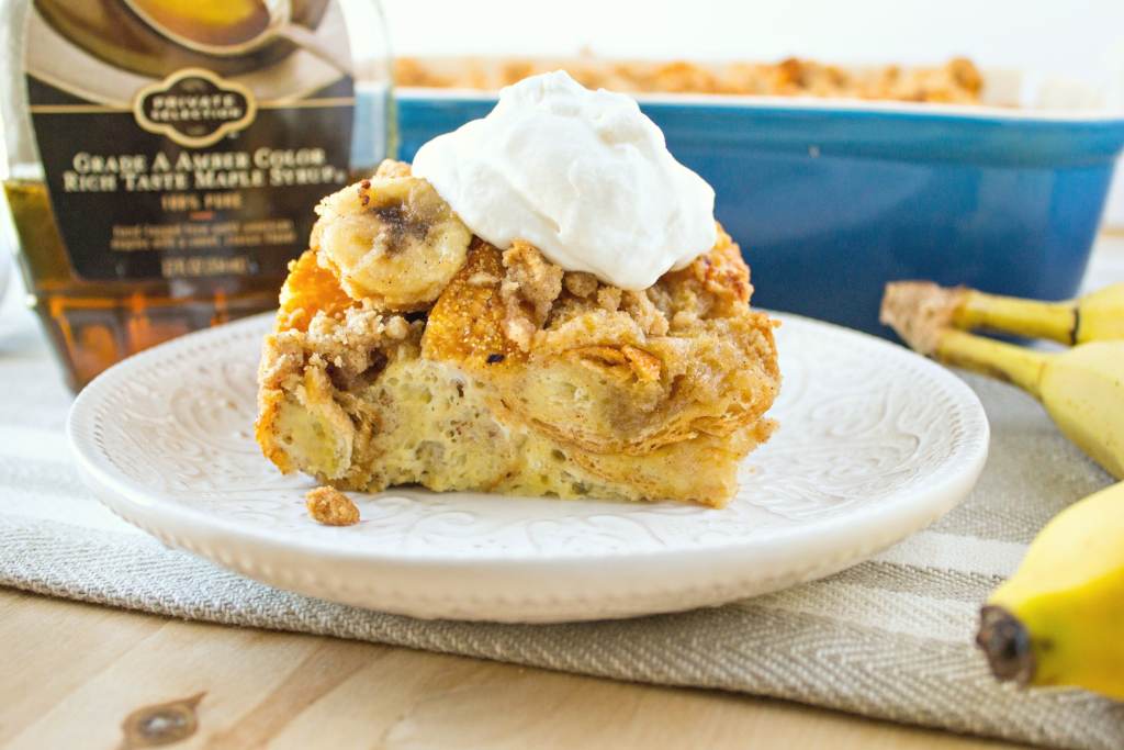 plate of bananas foster french toast casserole on a plate with a bottle of syrup and a baking dish with casserole in the background and a banana next to it, shot from the front