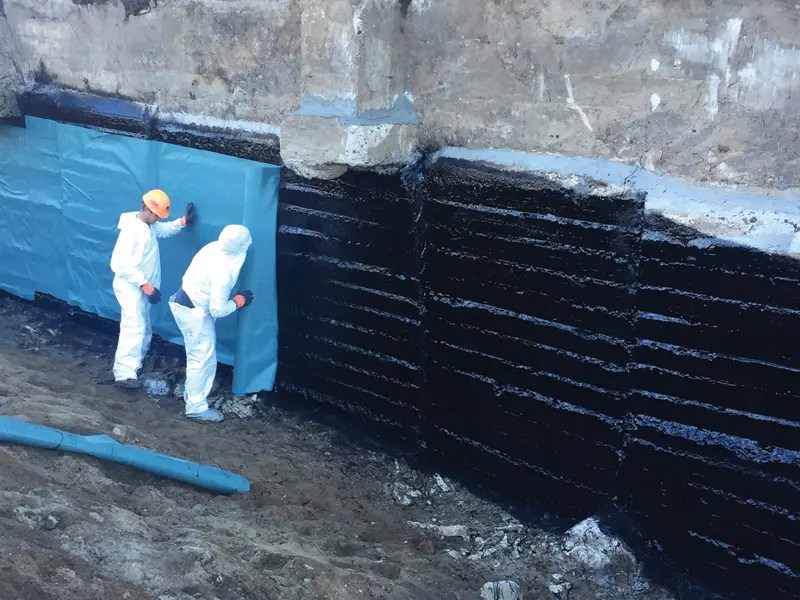 Two workers in white protective suits install a blue waterproof membrane against a black-coated wall in a construction site.