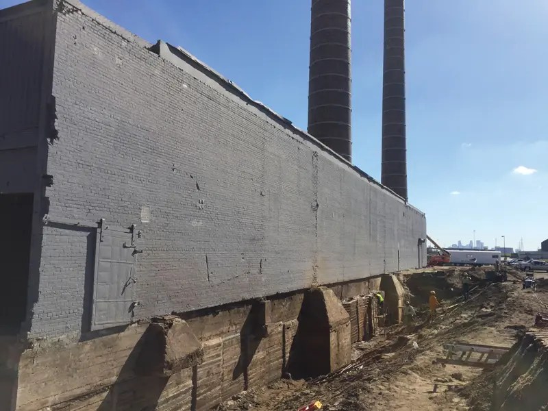Construction site featuring a gray brick building with two tall smokestacks, surrounded by workers and machinery under a blue sky.