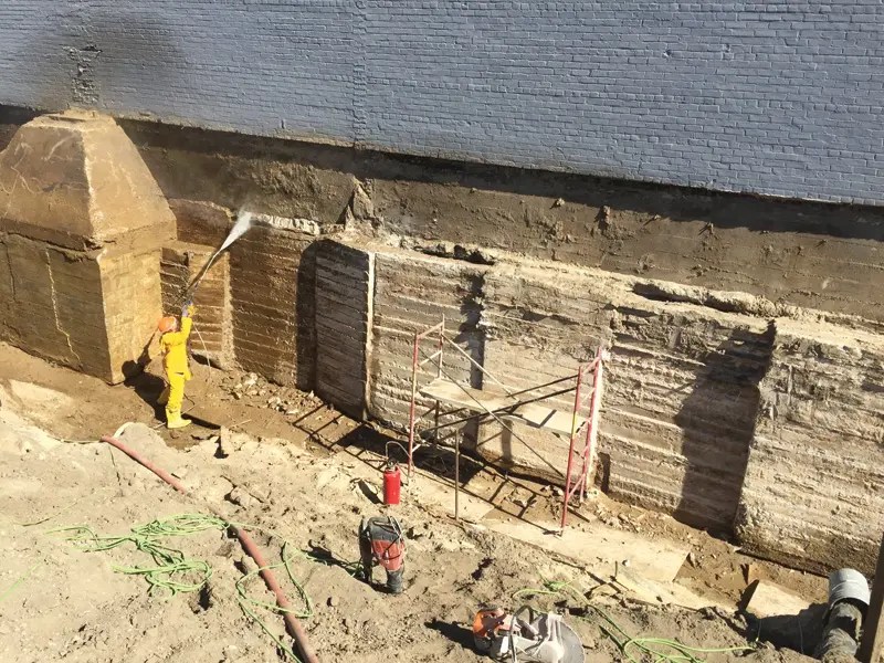 A worker in yellow overalls uses a high-pressure cleaner on a construction site, with scaffolding and exposed walls in the background.