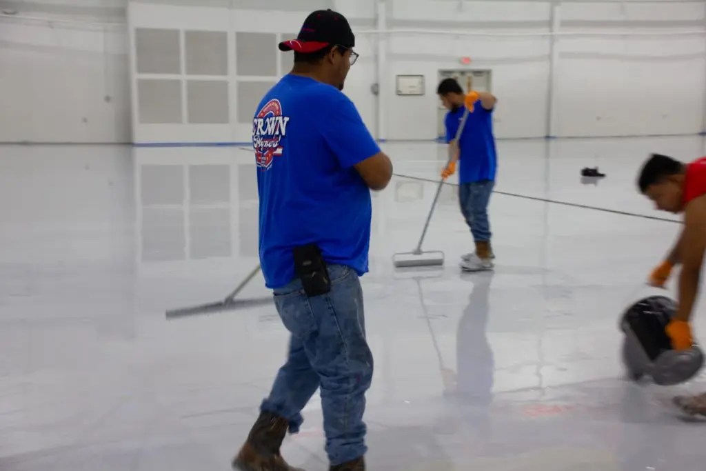 Workers in blue shirts and jeans apply a glossy finish to a large, empty indoor floor using rollers and buckets. Focused, active teamwork environment.