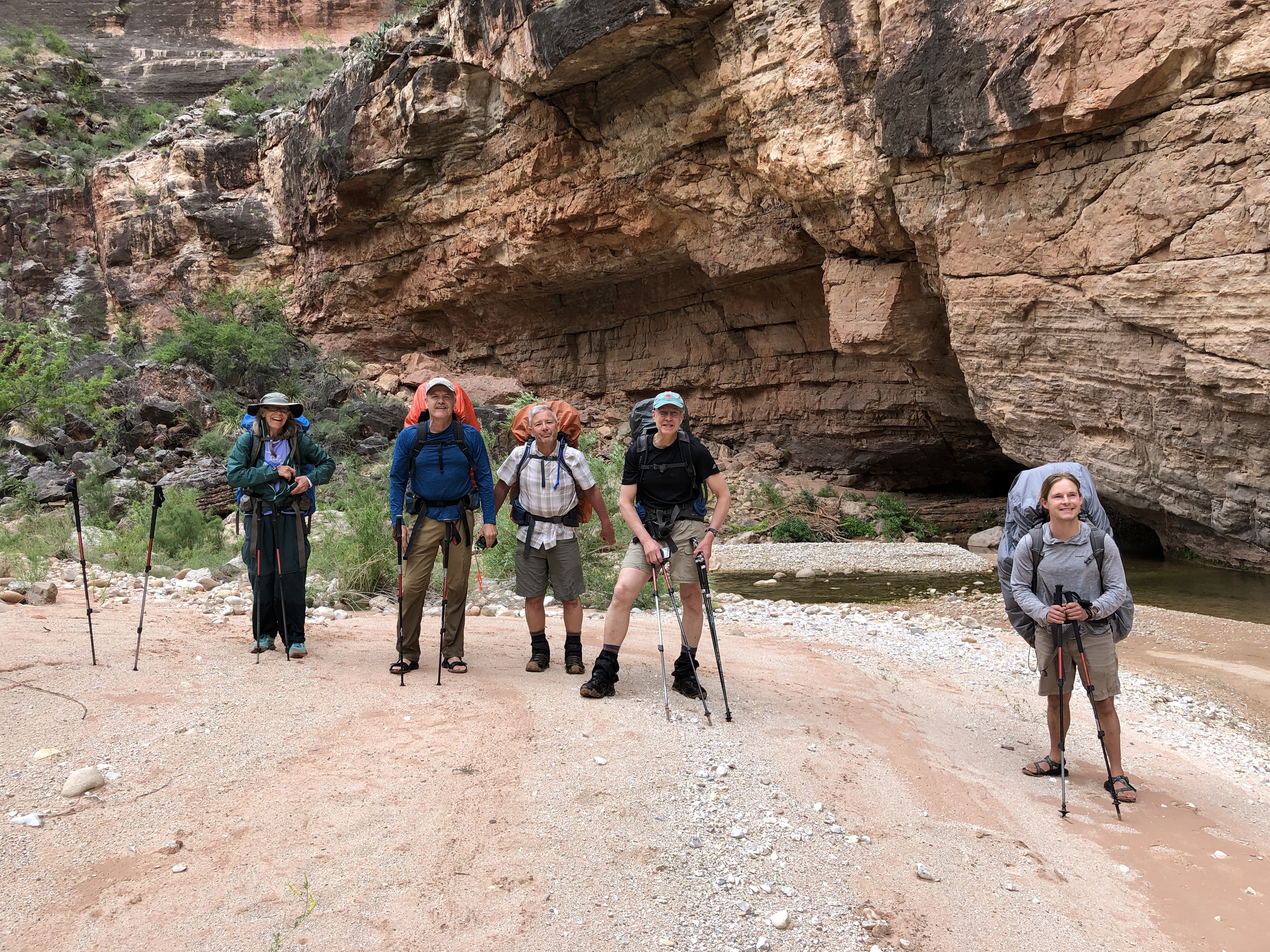 Five hikers standing beside a creek beneath a huge rock cliff.