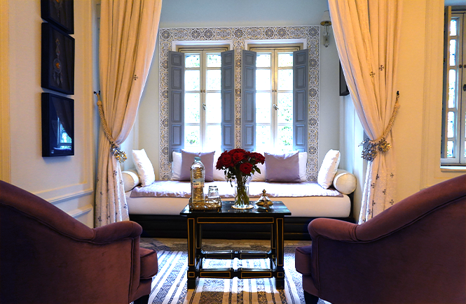 In the Aliyah suite at Riad Idra, Marrakech, Morocco, two mauve armchairs are placed in front of a black coffee table holding a vase of red roses, a silver tray, and a decorative glass bottle. Behind the table, sheer white curtains are pulled back to reveal a cozy daybed adorned with soft pillows, framed by intricate Moroccan tile work and large, traditional shuttered windows that let in natural light. The room exudes a tranquil, luxurious atmosphere with a mix of soft fabrics and detailed decor.
