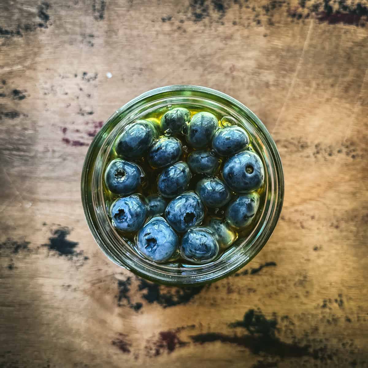A jar of blueberries floating in honey on a wood surface, top view.