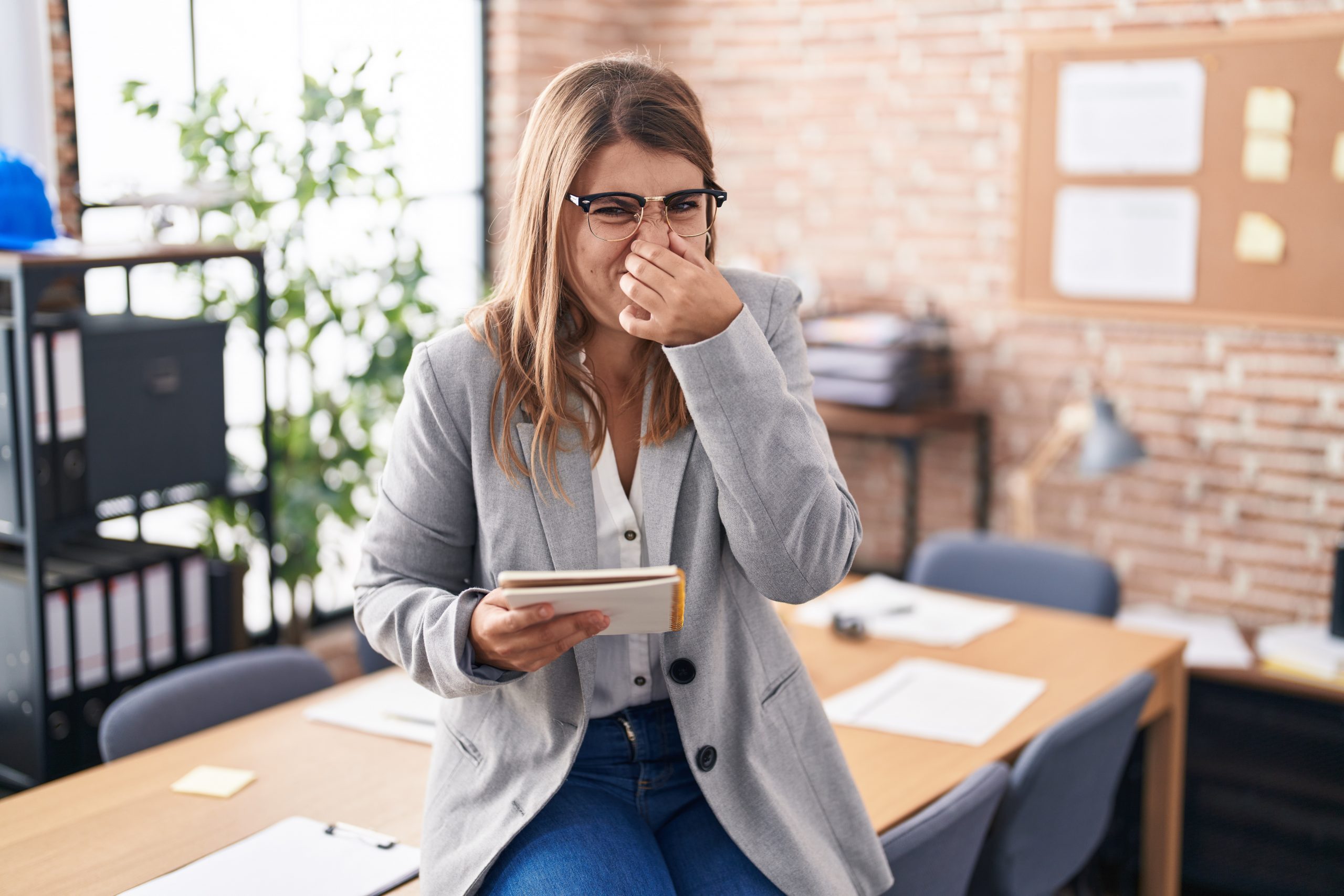Young woman working at the office wearing glasses smelling something