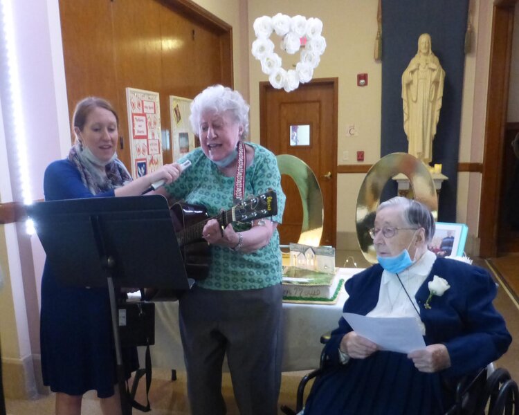 Sister Canice sits in a chair next to a pair of women playing guitar and singing.