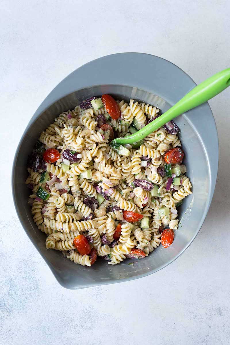 Using green spatula to combine rotini pasta, cherry tomatoes, kalamata olives and salad dressing in a gray bowl.