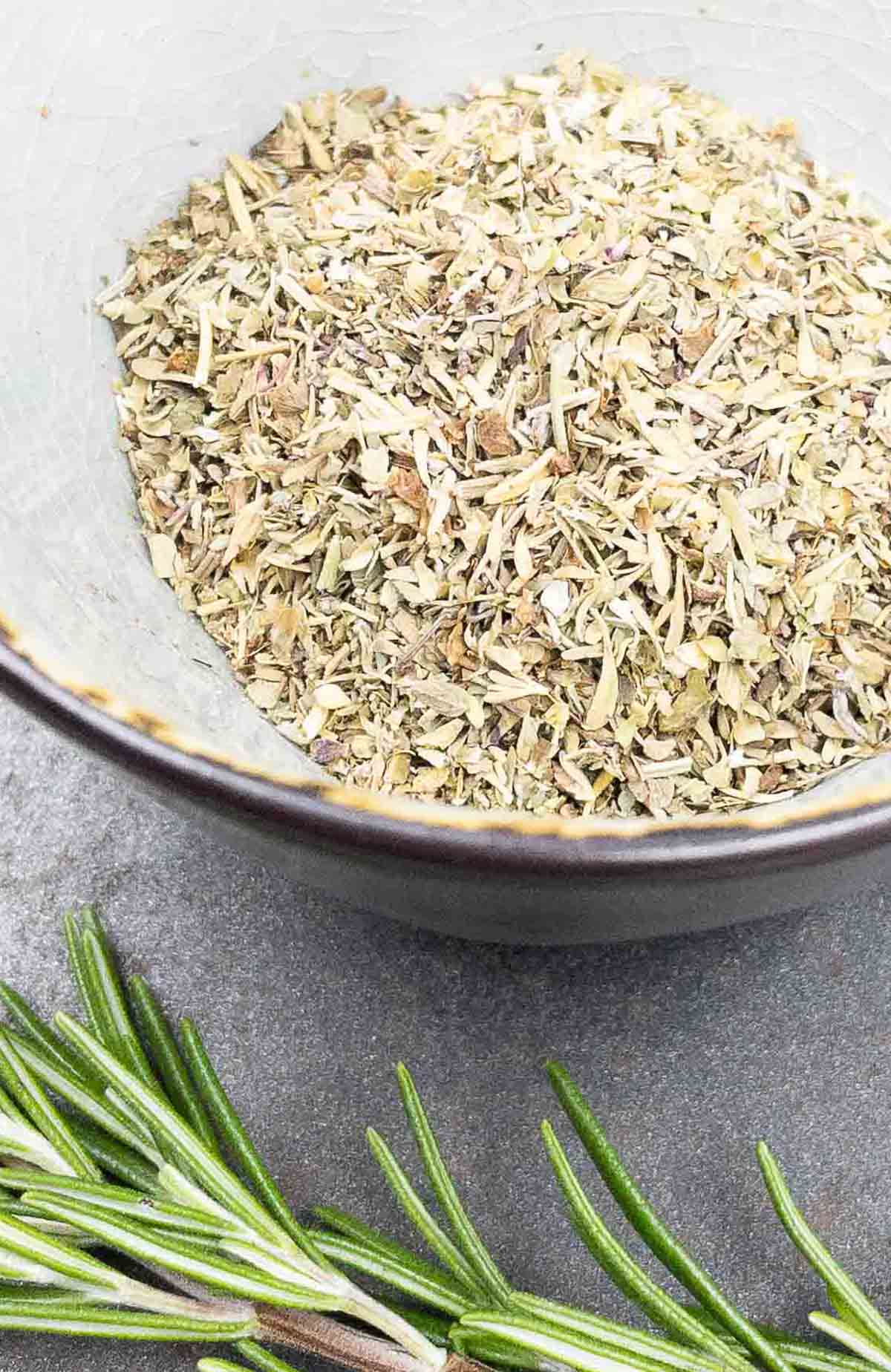 A bowl of homemade Italian seasoning and fresh rosemary sprig.