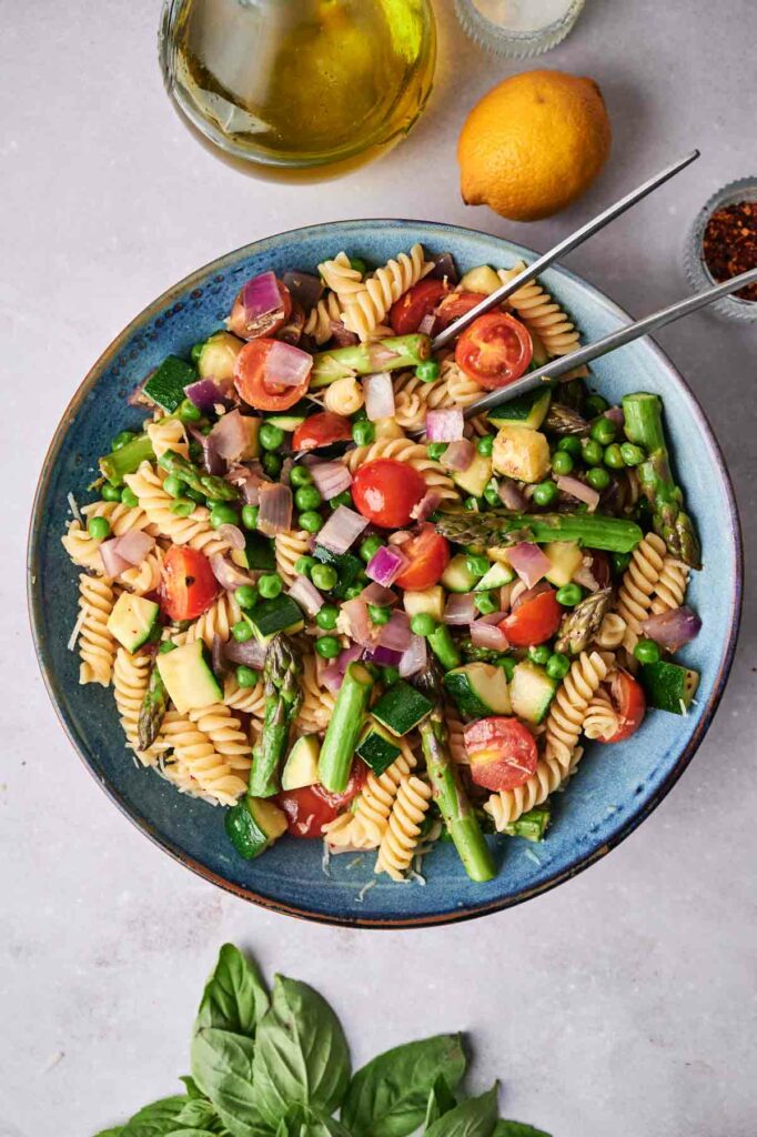 A plate of Pasta Primavera with tomatoes, asparagus, peas, and onions, served with a side dressing and lemon on a light background.