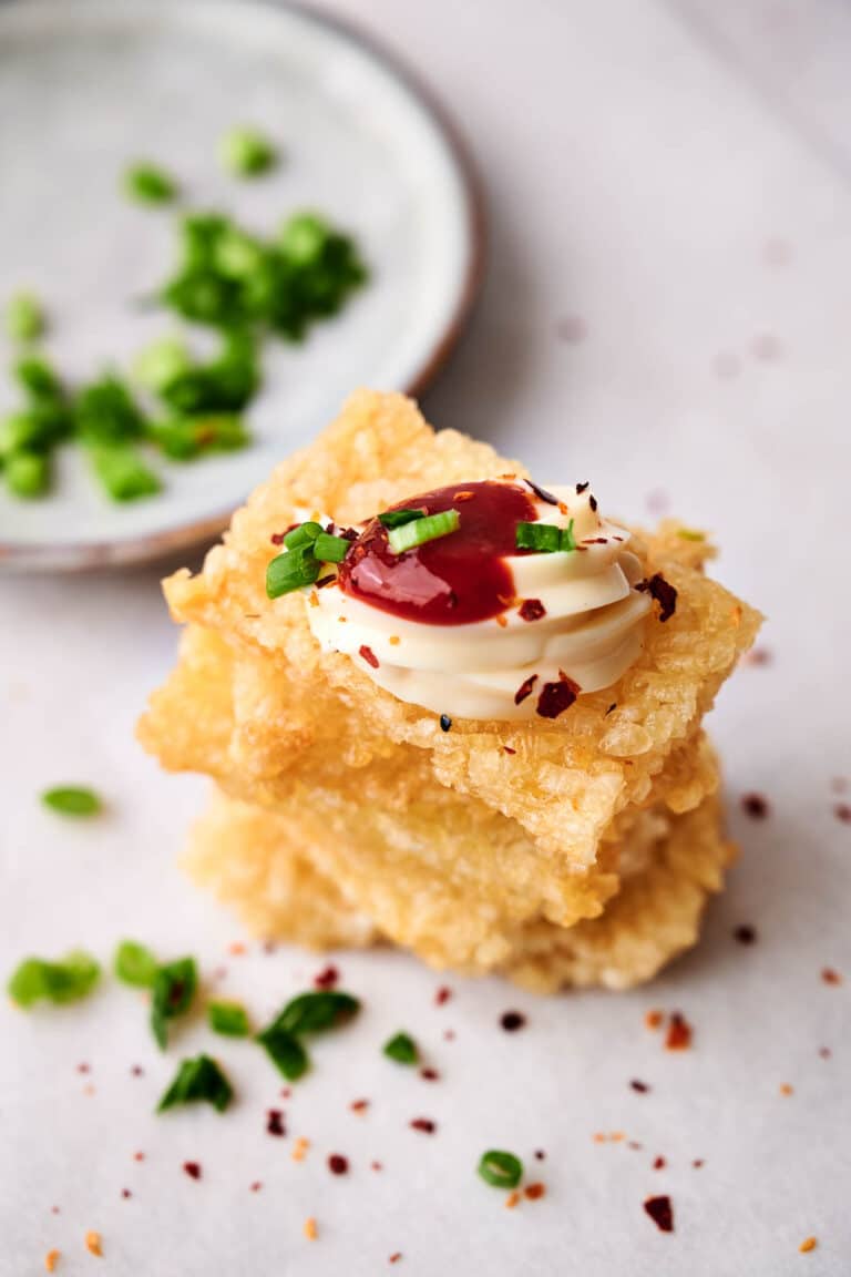A stack of crispy rice and fried tofu topped with mayonnaise, red sauce, and chopped green onions sits on a white surface, with a small dish of green onions in the background.