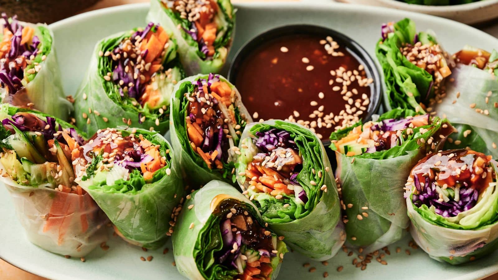 A plate of fresh vegetable spring rolls sliced in half, arranged around a dish of dipping sauce and sprinkled with sesame seeds.