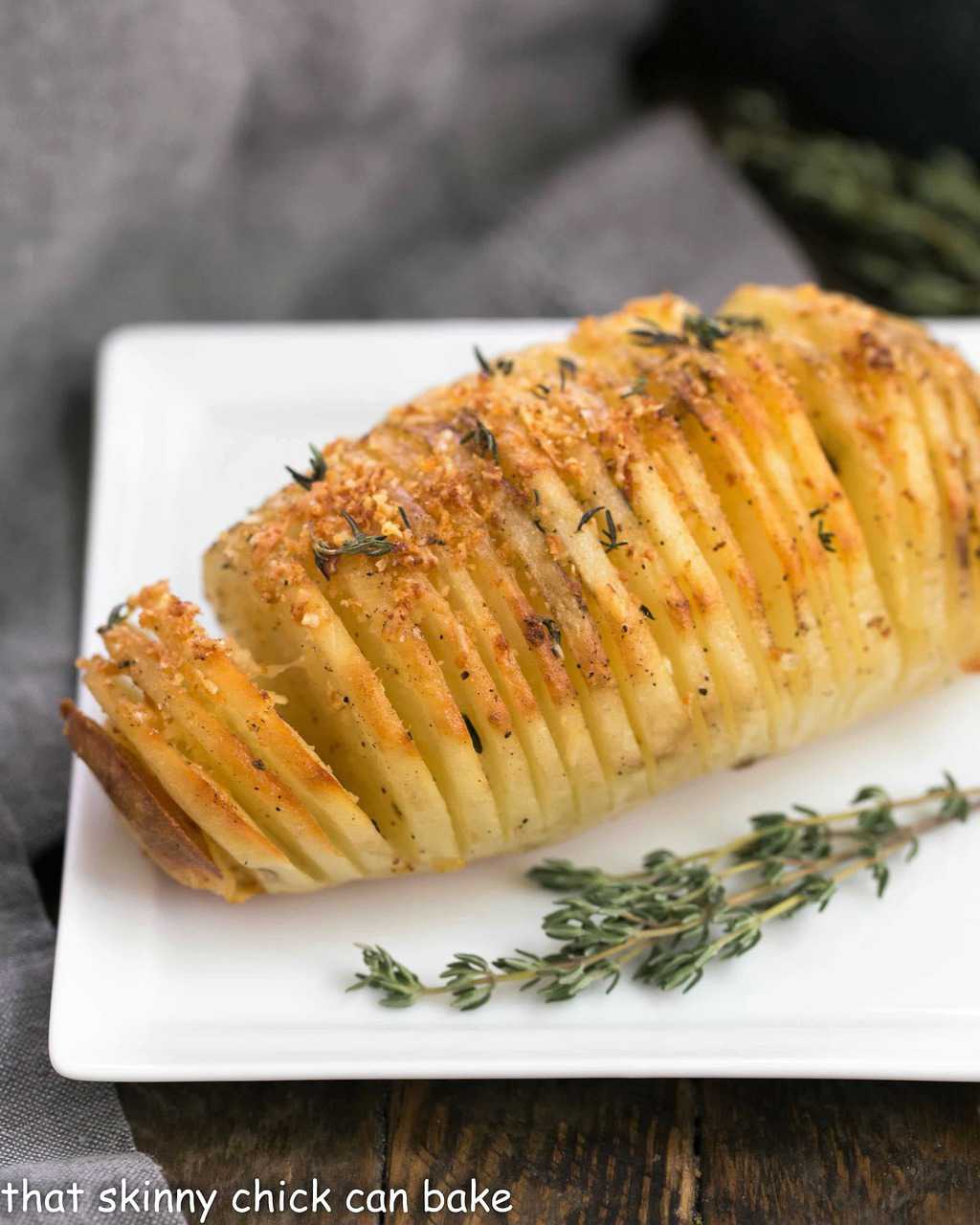 Hasselback Potatoes - Seasoned Fanned Potatoes that will delight your palate! Hasselback Potatoes on a square white plate with a sprig of thyme.