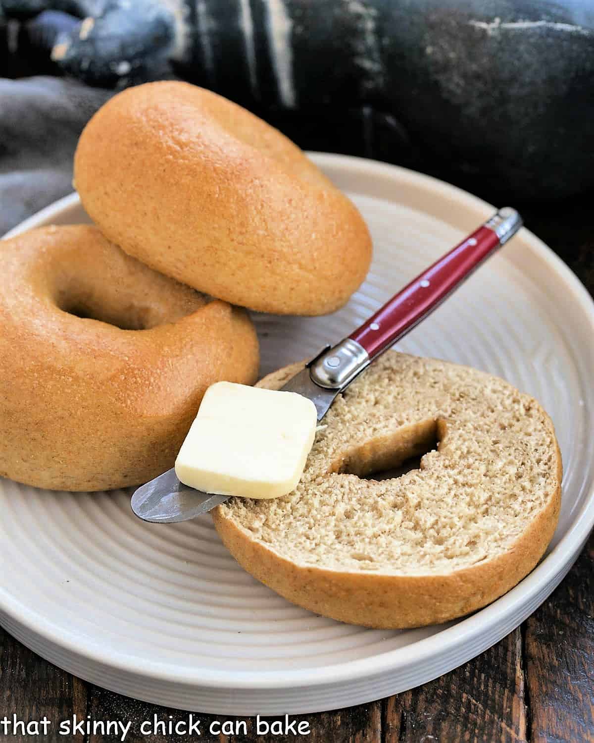 Whole Wheat Bagels - Dense, chewy and delicious and made in your own kitchen! 2 whole wheat bagels on a large white plate with one cut bagel topped with a red handle knife with a pat of butter.