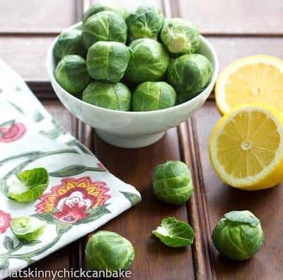 How about a raw Shaved Brussels Sprouts Salad with a lemony Dijon dressing? With Brussels sprouts being all the rage. This recipe is healthy and delicious! Brussels Sprouts in a small white bowl next to sliced lemons on a wooden table.