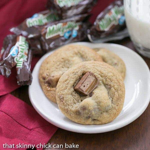 Milky Way Cookies are out of this world with a buttery, dark brown sugar base and chunks of candy bars!! These cookies are the perfect companion to a cold glass of milk. Three Milky Way Cookies on white plate surrounded by empty Milky Way wrappers.