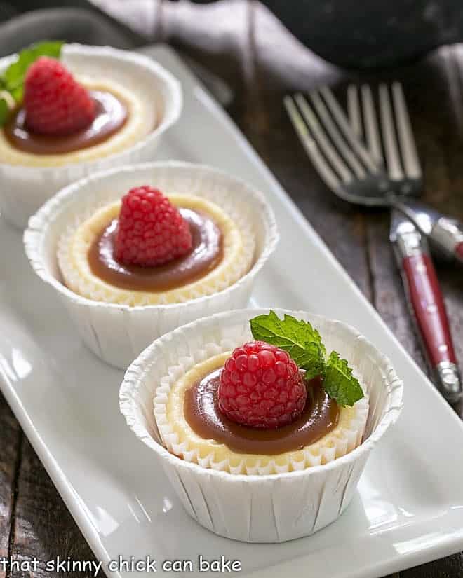 Caramel Topped Mini Cheesecakes - An exquisite combination of flavors and textures in these individual cheesecakes! Caramel Topped Mini Cheesecakes on a white rectangular tray garnished with raspberries.