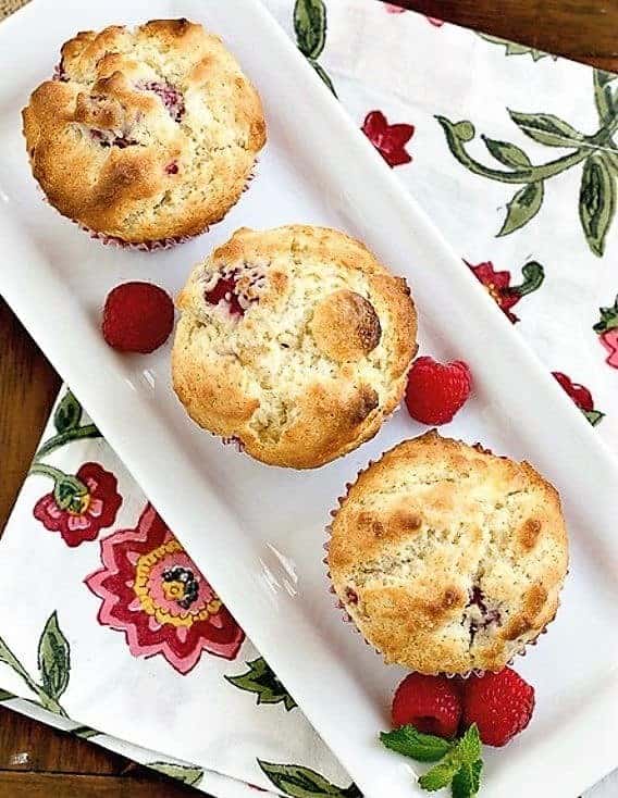 Spectacular Raspberry White Chocolate Muffins - sweet, tender and full of fresh berries and white chocolate Overhead view of 3 Raspberry White Chocolate Muffins on a rectangular tray with fresh raspberries and a sprig of mint.
