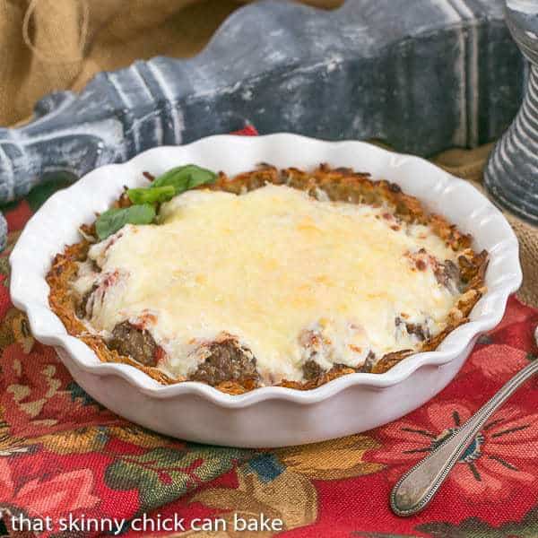 Italian Poutine Pie is a twist on the classic Canadian potato recipe. This Italian Poutine Pie recipe uses a hash brown crust, marinara and meatballs topped with gooey Mozzarella! Italian Poutine Pie in a white ceramic pie plate.