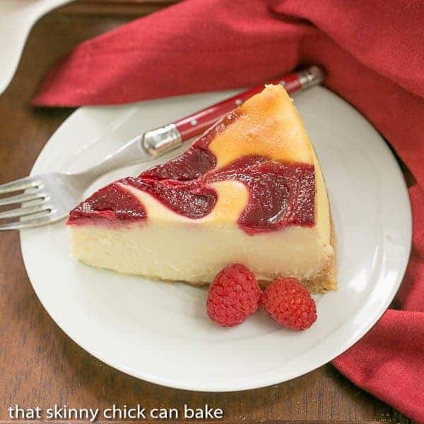 White Chocolate Raspberry Swirl Cheesecake - A luscious White Chocolate Cheesecake with a marbleized raspberry topping White Chocolate Raspberry Swirl Cheesecake slice on a white plate with fresh raspberries.
