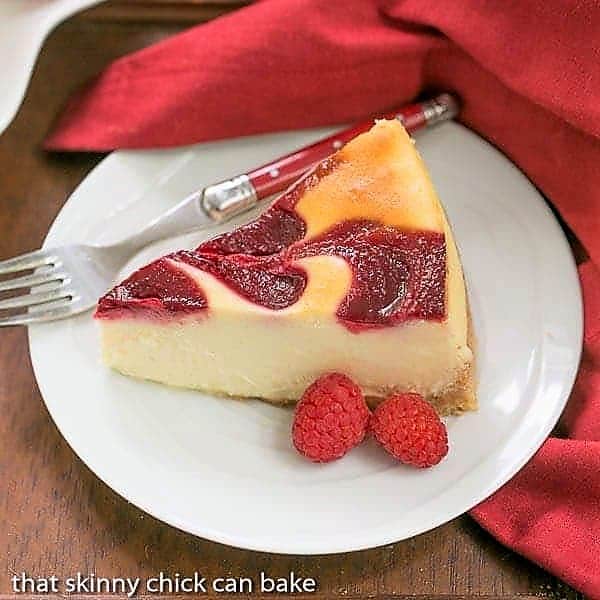 White Chocolate Raspberry Swirl Cheesecake - A luscious White Chocolate Cheesecake with a marbleized raspberry topping White Chocolate Raspberry Swirl Cheesecake slice on a round white plate with fresh raspberries and a red handle fork.