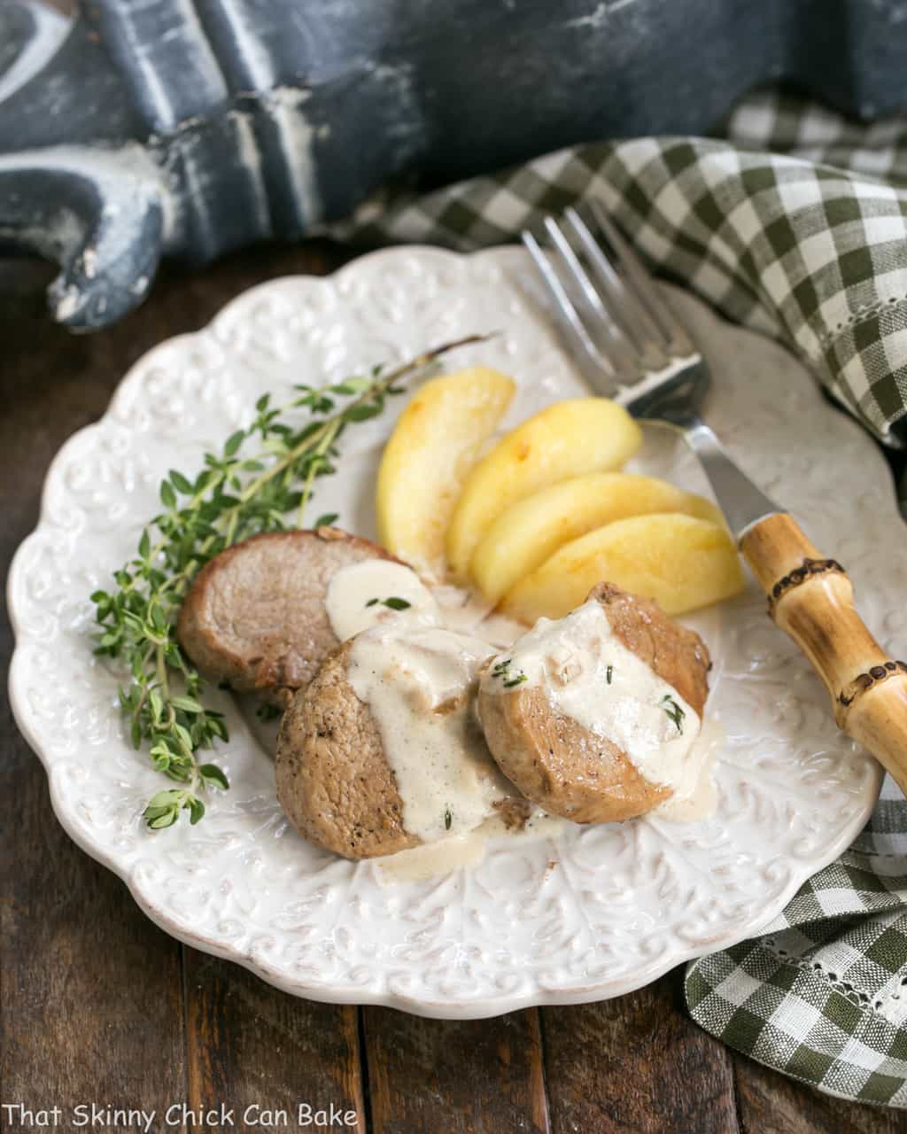 Apple Topped Pork Medallions with Calvados Cream Sauce - A gourmet entree that's easy enough for a weeknight! Apple Topped Pork Medallions with Calvados Cream Sauce on a dinner plate with fresh thyme and a bamboo handle fork.