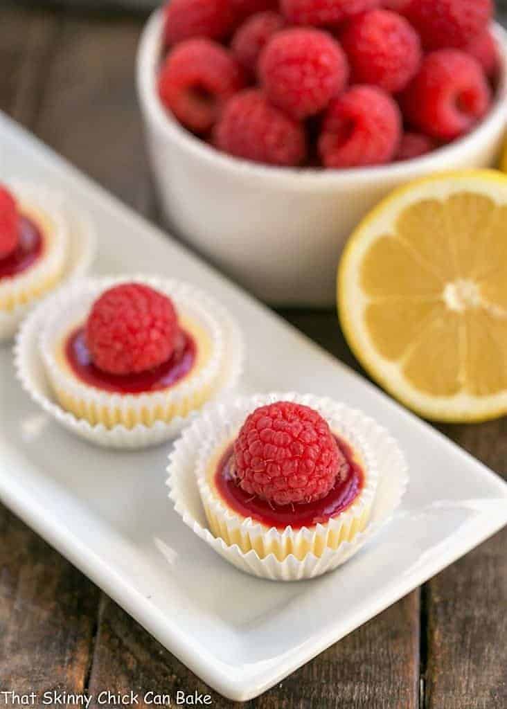 Mini Cheesecakes topped with a dab of raspberry sauce and a fresh raspberry, for an impressive, delectable dessert! Raspberry Topped Mini Cheesecakes on a white platter.
