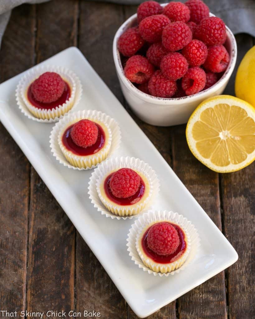 Raspberry Topped Mini Cheesecakes - Creamy mini cheesecakes topped with raspberry sauce and a plump ripe raspberry! Overhead view of a white dessert platter with bite sized cheesecakes topped with raspberry sauce and fresh raspberries