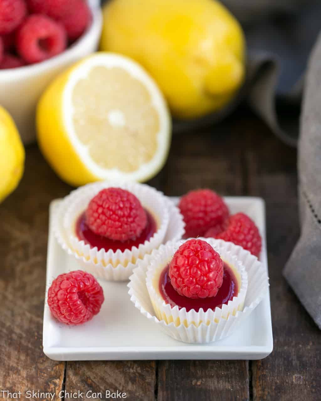 Raspberry Topped Mini Cheesecakes - Creamy mini cheesecakes topped with raspberry sauce and a plump ripe raspberry! Two raspberry topped cheesecake bites on a small white plate with raspberry garnishes.