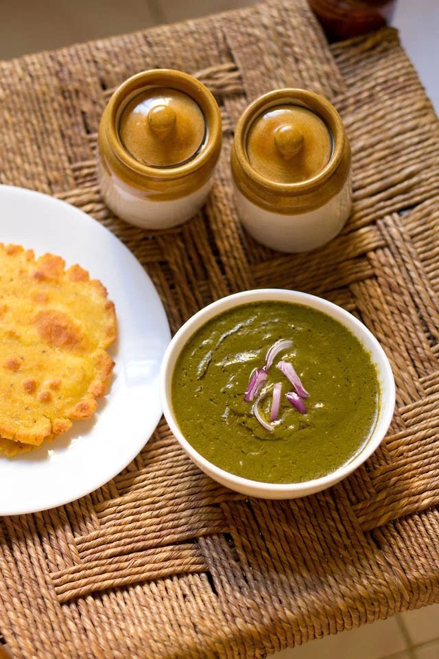 sarson ka saag sarson ka saag in a white bowl on a rattan table with a white plate of makki ki roti to the side.