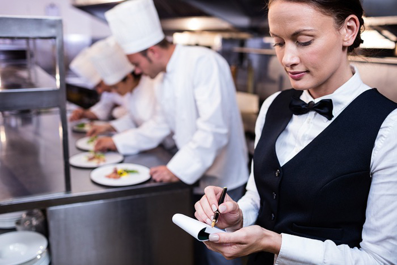 Waitress with notepad in commercial kitchen and chefs preparing Your