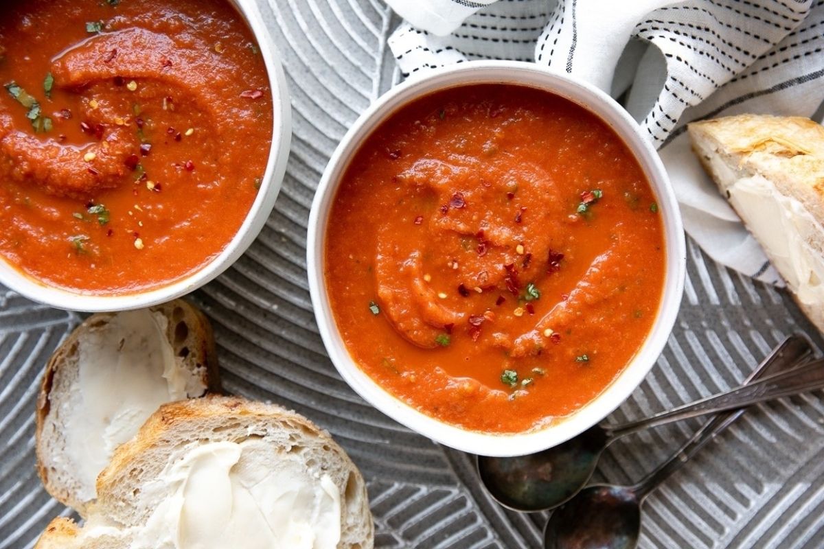 Two bowls of tomato based soup next to a piece of bread.