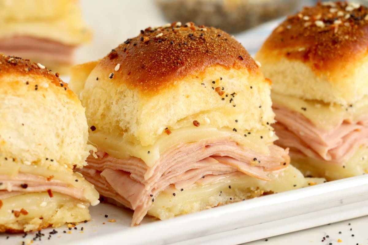 Sliced ham and cheese slider recipes with a poppy seed topping on a white plate.