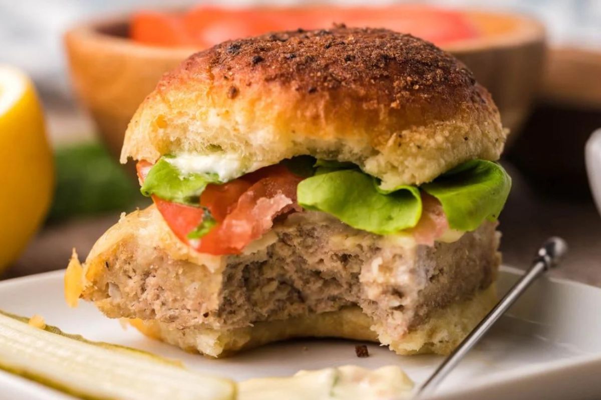 A turkey burger slider with lettuce, tomato, and mayonnaise on a plate, partially eaten, with a pickle on the side.