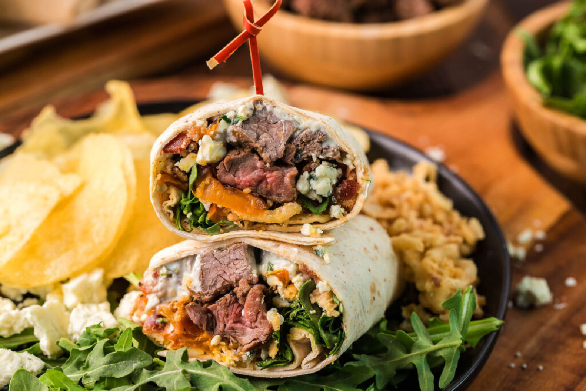 Steak and blue cheese wrap cut in half, served with chips and rice on a wooden table.