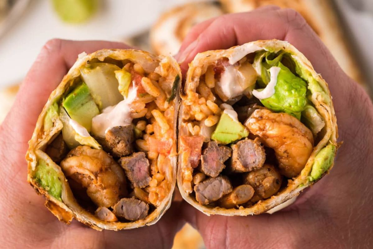 Two hands holding a halved burrito filled with shrimp, rice, beans, avocado, and vegetables, viewed up close.