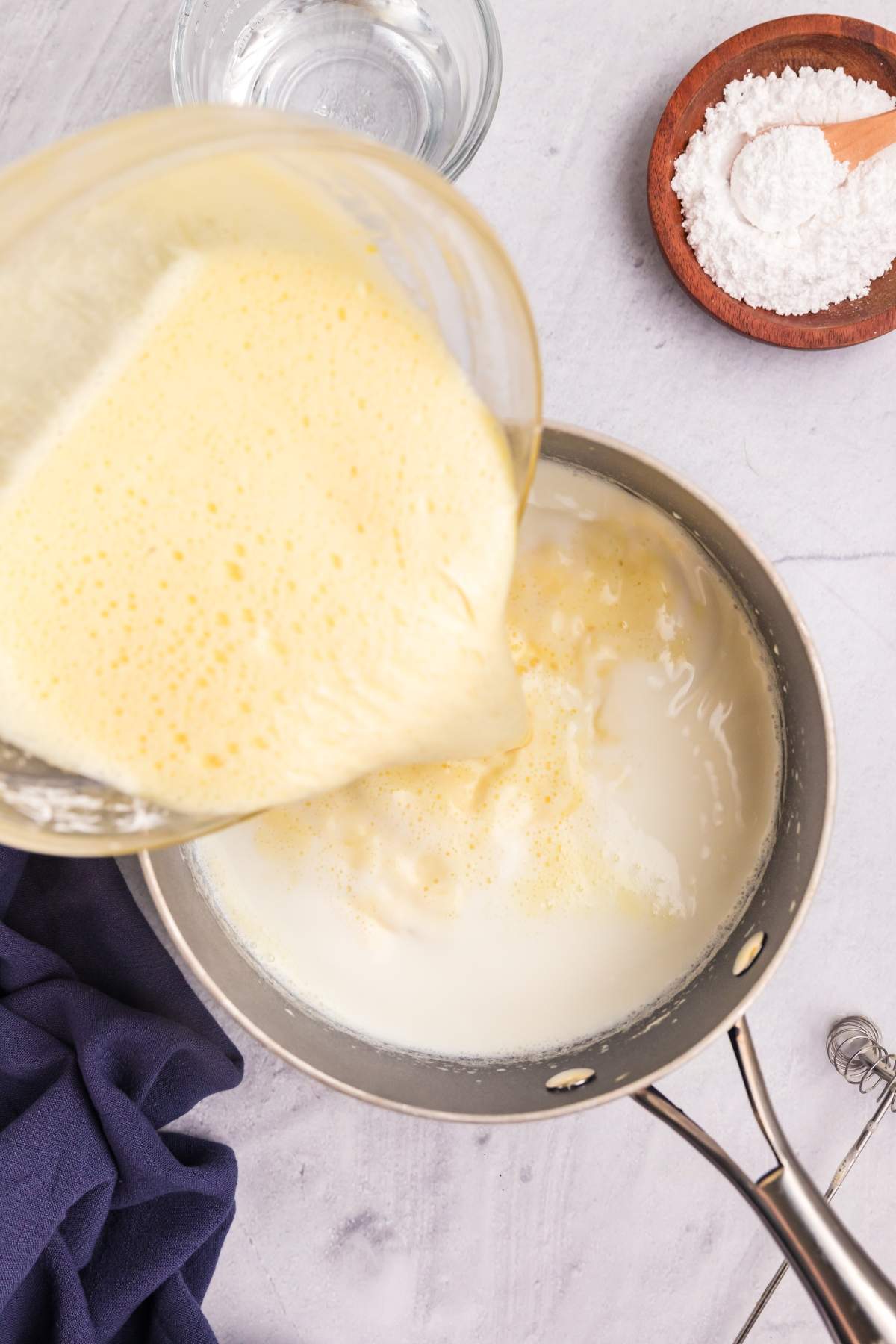 A mixture is being poured into a saucepan containing milk on a stovetop, with a bowl of flour and a glass of water nearby, preparing the base for a delicious torta della nonna.