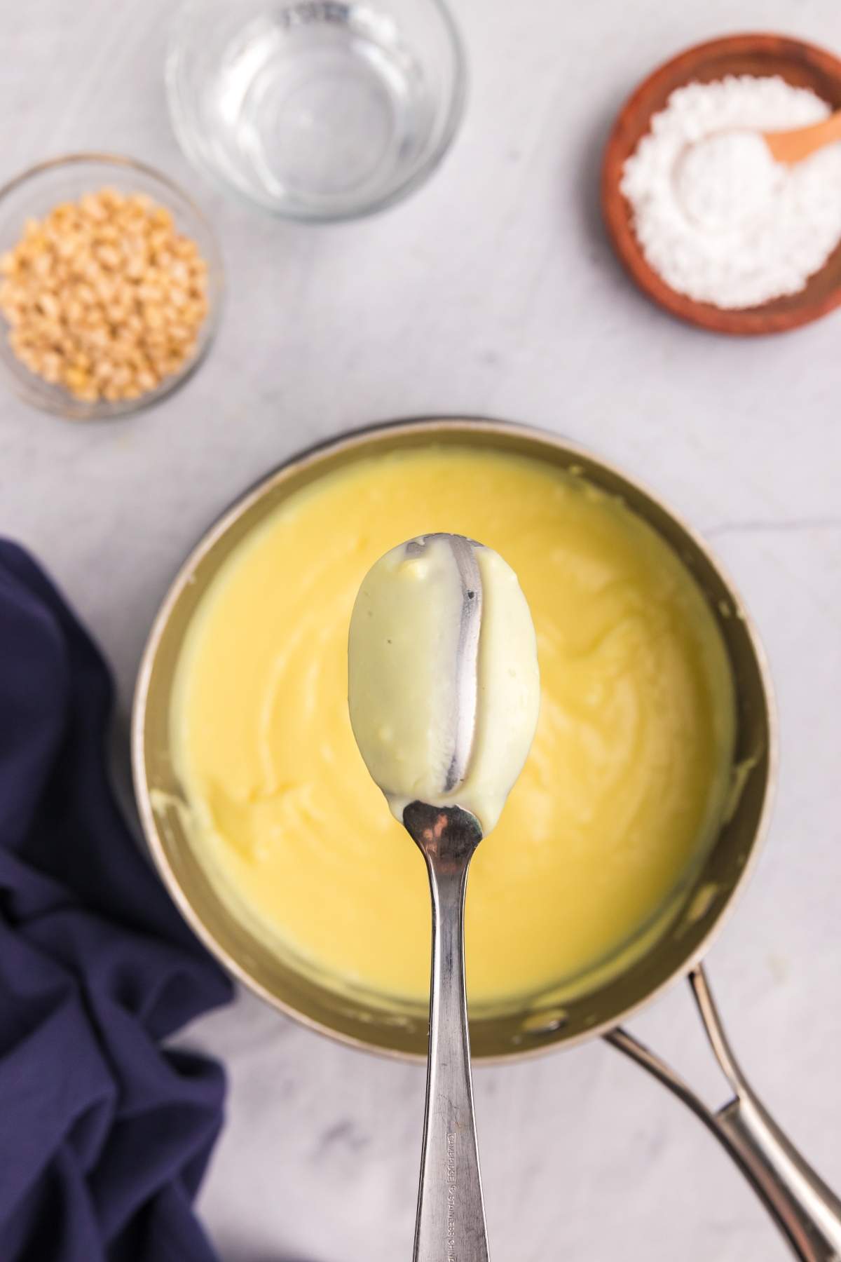 A metal spoon with a thick, creamy yellow sauce is held above a pot of the same luscious mixture, reminiscent of the filling for torta della nonna. In the background, small bowls containing popcorn kernels and flour can be seen, hinting at ingredients ready for other culinary creations.