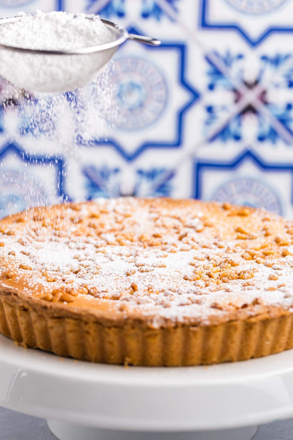 A torta della nonna on a white stand is being dusted with powdered sugar using a metal sieve. The background features blue and white patterned tiles.