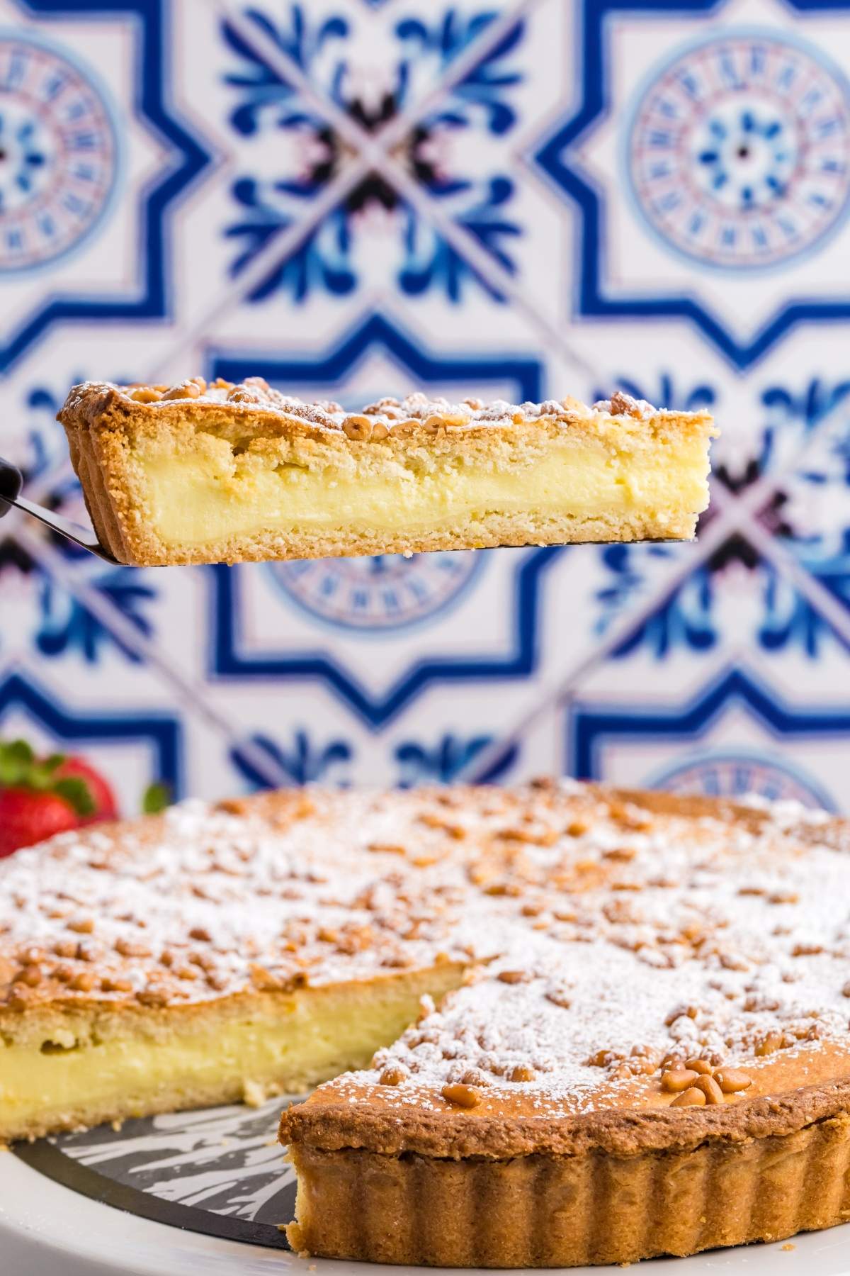 A slice of powdered sugar-topped torta della nonna custard pie is being lifted from a pie with a patterned blue and white tile background.