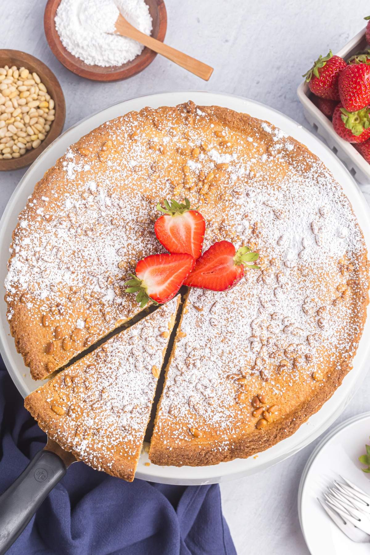 A torta della nonna recipe dusted with powdered sugar, topped with sliced strawberries, and partially sliced. Pine nuts and a bowl of strawberries are nearby.