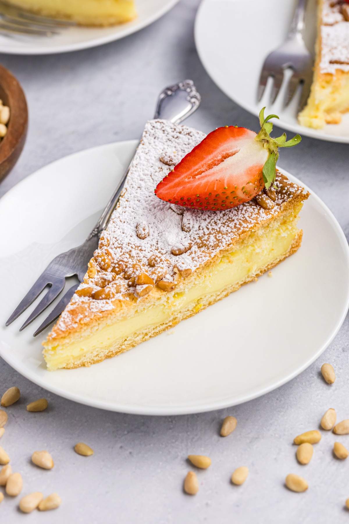 A slice of Italian torta della nonna cake topped with a halved strawberry and powdered sugar on a white plate, with a fork beside the slice. Scattered pine nuts are visible on the table around the plate.